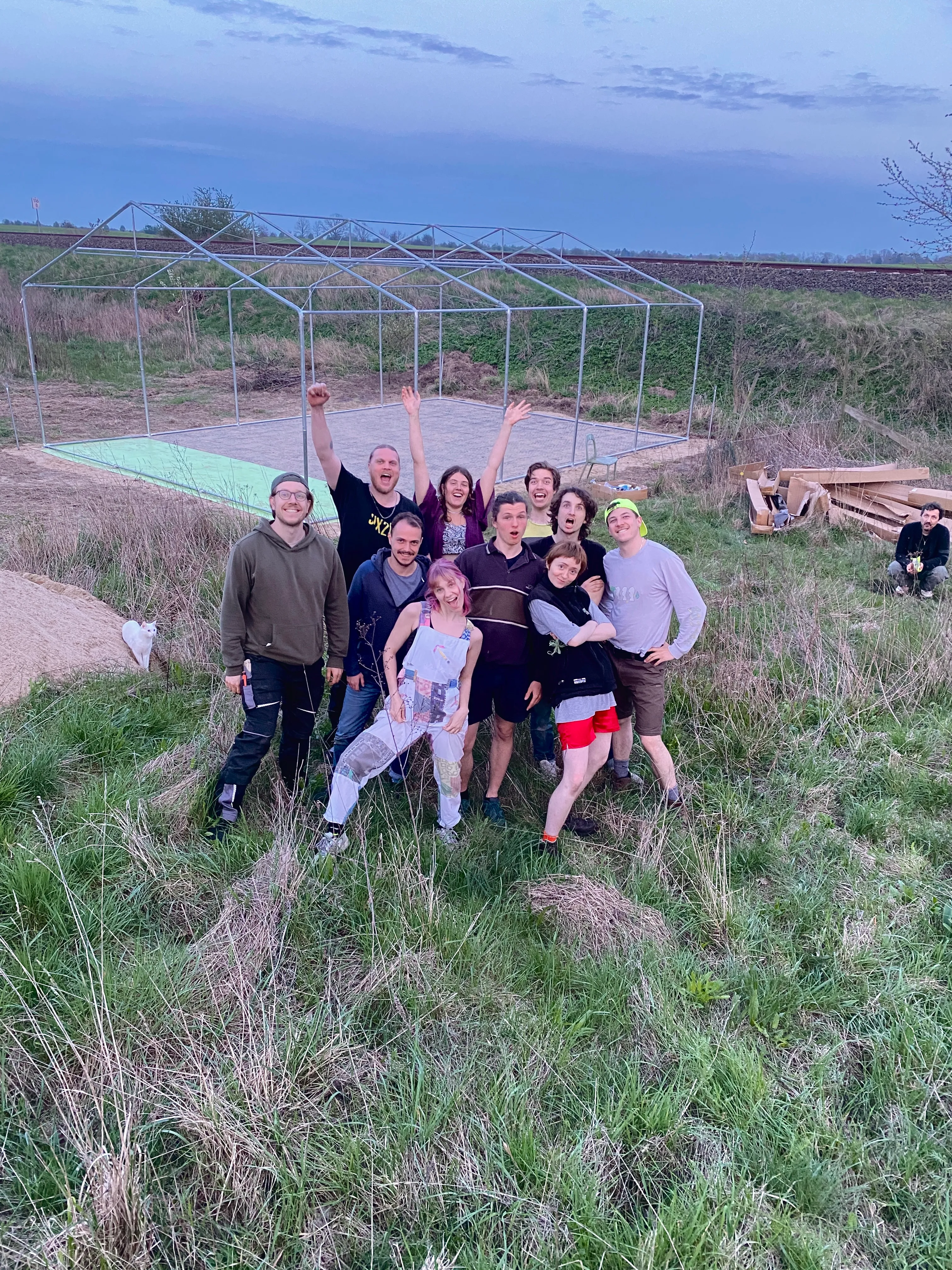 Eine Gruppe junger Menschen steht zusammen auf einer Wiese vor dem Gerüst eines Partyzelts. Sie ziehen Grimassen und stehen in witzigen und coolen Posen für die Kamera. 